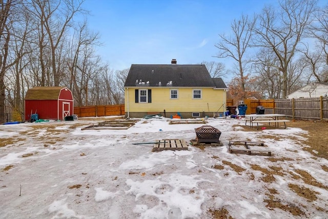 snow covered rear of property with an outdoor fire pit, a fenced backyard, a chimney, an outdoor structure, and a shed