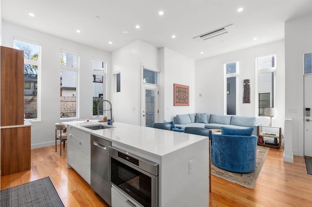 kitchen featuring stainless steel dishwasher, a kitchen island with sink, sink, light hardwood / wood-style flooring, and white cabinets