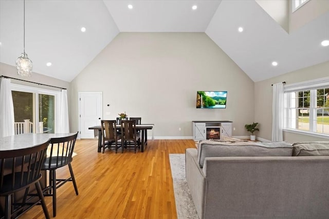 living room featuring high vaulted ceiling, light wood-style flooring, a warm lit fireplace, recessed lighting, and baseboards