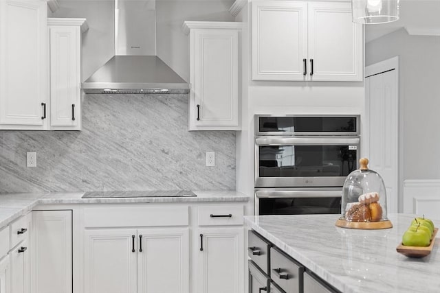 kitchen featuring backsplash, wall chimney range hood, stainless steel double oven, light stone counters, and white cabinetry