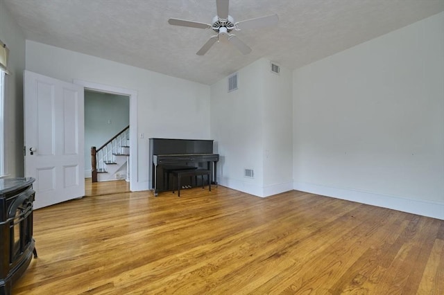 unfurnished living room with a wood stove, light wood finished floors, stairs, a ceiling fan, and baseboards