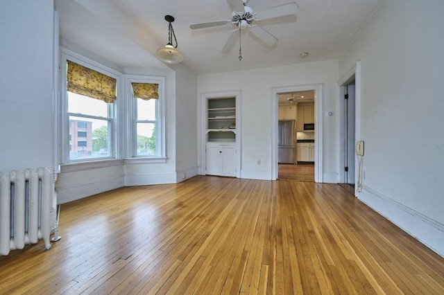 empty room with radiator heating unit, light wood-type flooring, ceiling fan, built in shelves, and baseboards