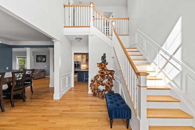 stairs with wood finished floors, ornate columns, a decorative wall, crown molding, and a towering ceiling