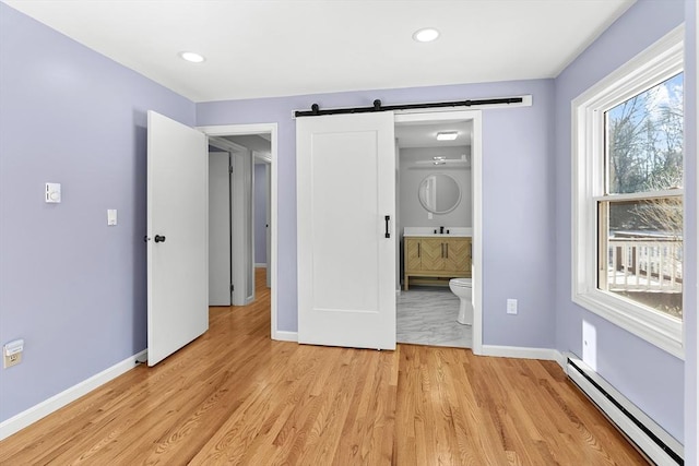 unfurnished bedroom featuring light wood-type flooring, a barn door, ensuite bath, and a baseboard heating unit