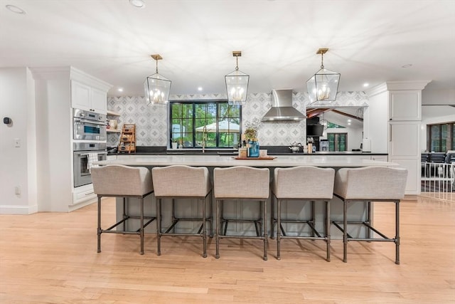 4 Rockwood Ln, Londonderry, NH 03053 - kitchen featuring wall chimney exhaust hood, white cabinetry, double oven, a breakfast bar area, and light wood-style floors