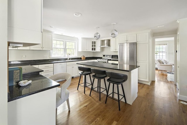 kitchen featuring a breakfast bar, wall chimney exhaust hood, dark countertops, white cabinets, and appliances with stainless steel finishes