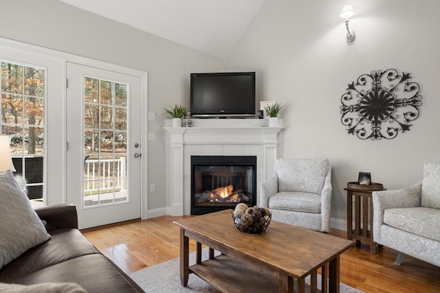 living area featuring baseboards, light wood-style flooring, a tiled fireplace, and vaulted ceiling