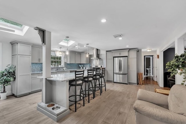 kitchen featuring gray cabinetry, wall chimney exhaust hood, stainless steel refrigerator, and a skylight