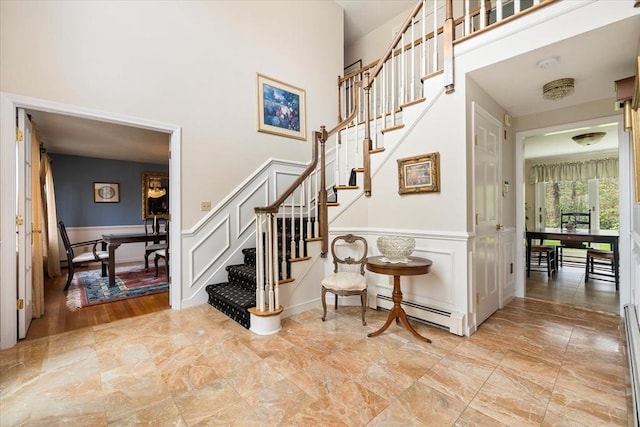 stairs featuring a towering ceiling, a wainscoted wall, baseboard heating, and a decorative wall