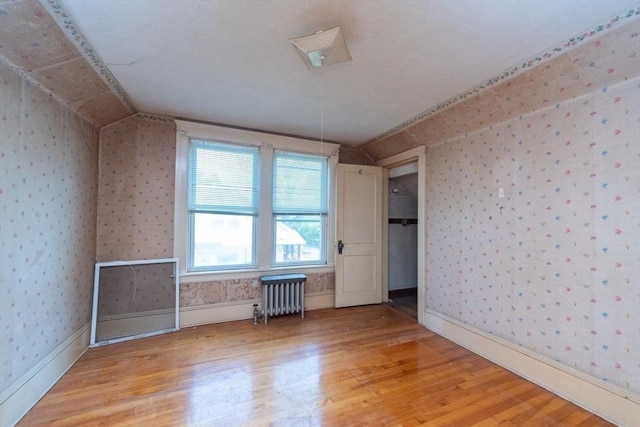 unfurnished bedroom with radiator, light wood-style floors, wallpapered walls, lofted ceiling, and baseboards