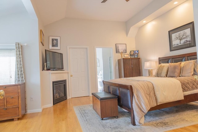 bedroom with baseboards, connected bathroom, a glass covered fireplace, lofted ceiling, and light wood-style floors