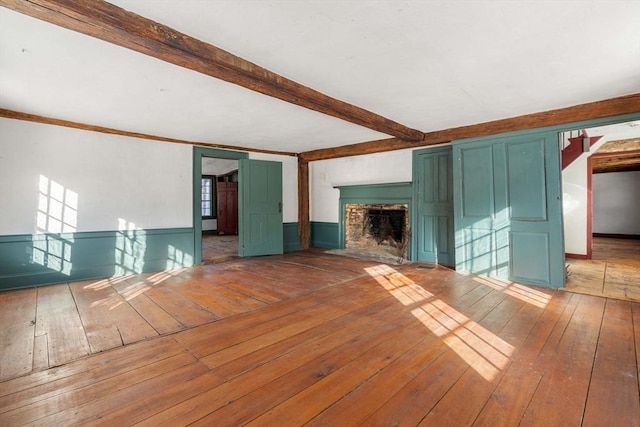 unfurnished living room with a wainscoted wall, beamed ceiling, wood-type flooring, and a brick fireplace