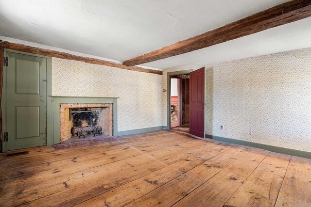 unfurnished living room featuring baseboards, light wood finished floors, beamed ceiling, a wood stove, and wallpapered walls