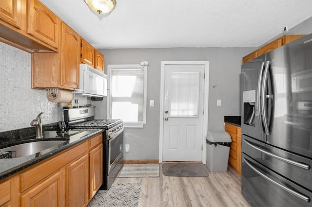 kitchen featuring light wood finished floors, decorative backsplash, appliances with stainless steel finishes, dark stone countertops, and a sink