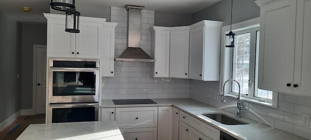 kitchen with wall chimney range hood, black appliances, sink, hanging light fixtures, and white cabinets