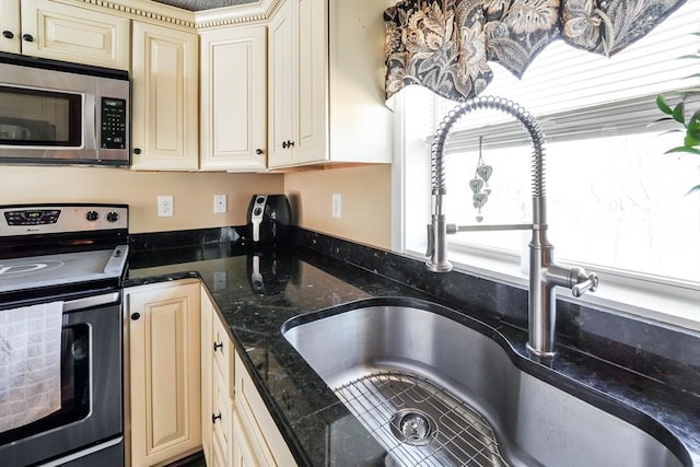 kitchen with dark stone countertops, cream cabinetry, a sink, and appliances with stainless steel finishes