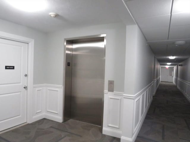 corridor with elevator, wainscoting, and dark carpet