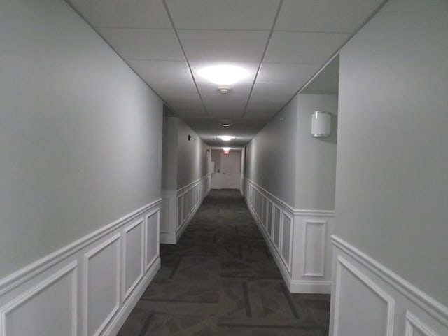 hallway featuring a drop ceiling, a decorative wall, wainscoting, and dark colored carpet