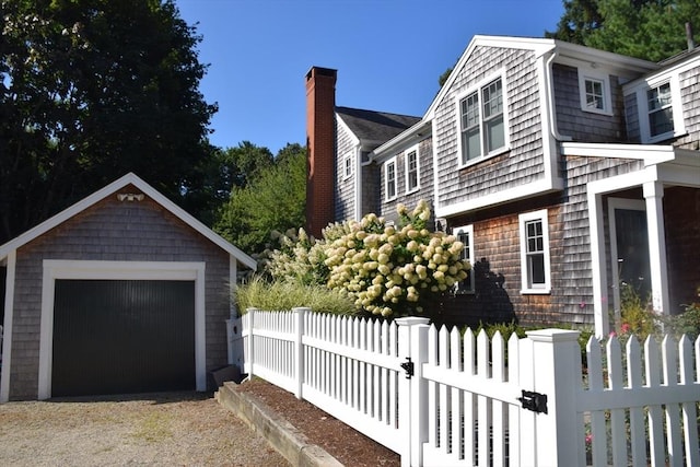 view of side of property with a detached garage, gravel driveway, a fenced front yard, a chimney, and an outbuilding