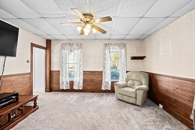 living area with ceiling fan, carpet flooring, wooden walls, and a drop ceiling