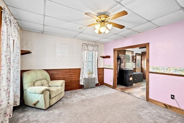 living area featuring wood walls, light colored carpet, a wood stove, ceiling fan, and a drop ceiling