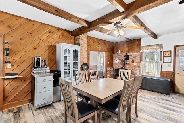 dining room with a wood stove, ceiling fan, beam ceiling, wooden walls, and light wood-type flooring