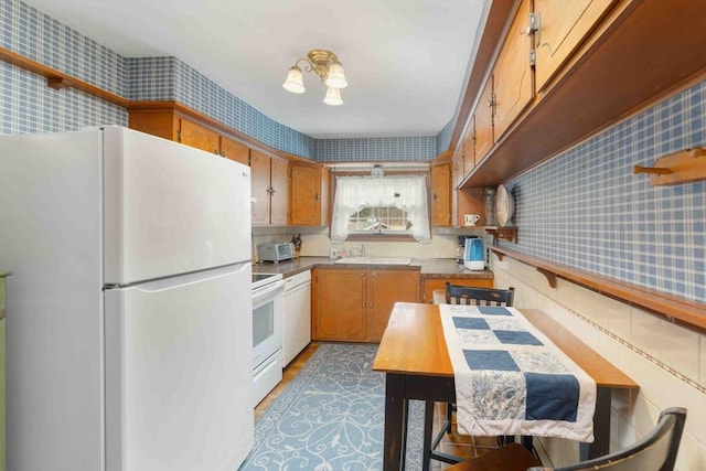 kitchen with a wainscoted wall, white appliances, a sink, brown cabinetry, and wallpapered walls