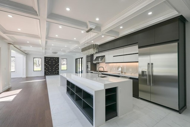 kitchen with appliances with stainless steel finishes, coffered ceiling, exhaust hood, a center island with sink, and hanging light fixtures