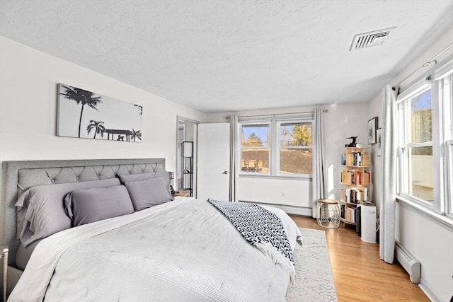 bedroom featuring a baseboard heating unit, light wood-style floors, visible vents, and a textured ceiling