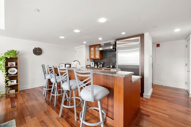 kitchen with wall chimney range hood, decorative backsplash, stainless steel built in fridge, and wood finished floors
