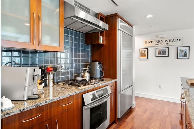 kitchen with brown cabinetry, decorative backsplash, wall chimney exhaust hood, light stone counters, and appliances with stainless steel finishes