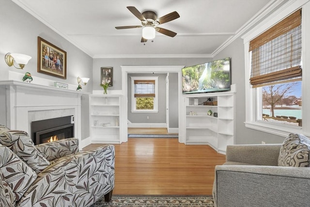 living area with baseboards, a ceiling fan, a glass covered fireplace, ornamental molding, and wood finished floors