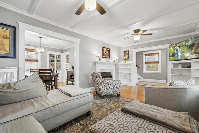 living area featuring ceiling fan with notable chandelier, a fireplace, wood finished floors, baseboards, and crown molding