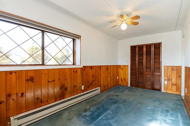 unfurnished bedroom featuring wooden walls, a wainscoted wall, carpet, baseboard heating, and a closet