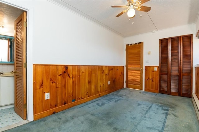 carpeted empty room featuring wainscoting, ceiling fan, ornamental molding, wood walls, and a baseboard heating unit