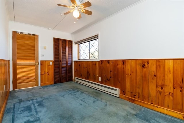 carpeted spare room featuring ceiling fan, wood walls, baseboard heating, wainscoting, and crown molding