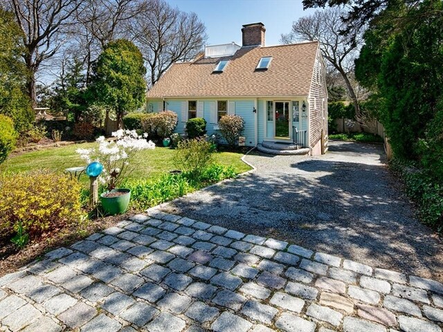 cape cod-style house with a shingled roof, a front yard, driveway, and a chimney