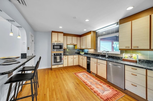 kitchen with appliances with stainless steel finishes, a sink, light brown cabinetry, tasteful backsplash, and dark stone counters