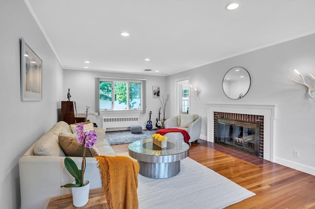 living area with radiator, ornamental molding, a brick fireplace, recessed lighting, and wood finished floors