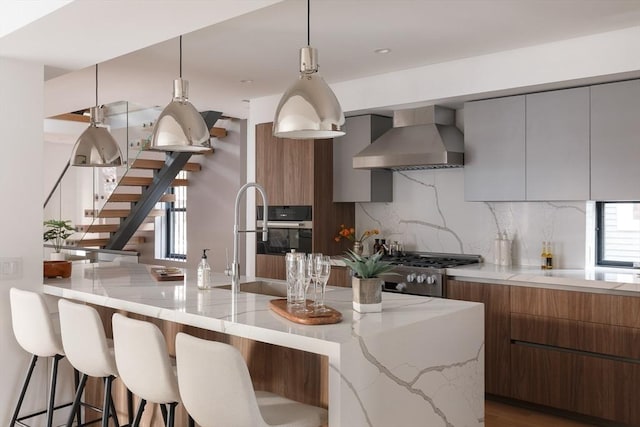 kitchen with sink, wall chimney range hood, light stone counters, backsplash, and range