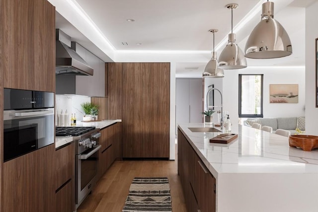 kitchen with stainless steel appliances, wall chimney range hood, light stone counters, pendant lighting, and light hardwood / wood-style floors