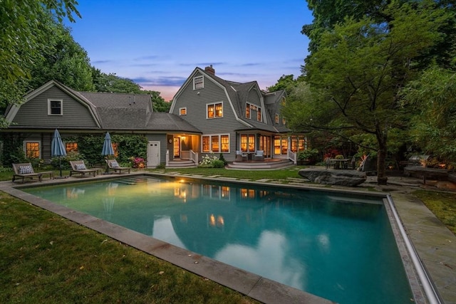 back of house at dusk with a chimney, a patio area, a gambrel roof, and an outdoor pool