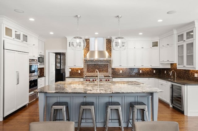 kitchen featuring wall chimney range hood, a kitchen island with sink, glass insert cabinets, and paneled refrigerator