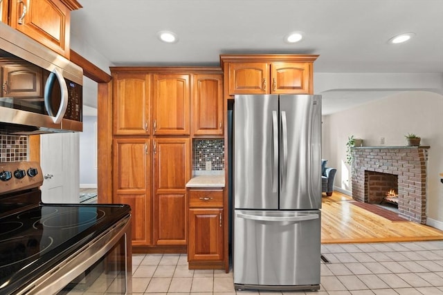 kitchen with light tile patterned floors, stainless steel appliances, brown cabinetry, a brick fireplace, and tasteful backsplash