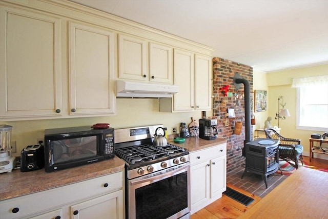kitchen featuring cream cabinets, a wood stove, under cabinet range hood, and stainless steel range with gas cooktop