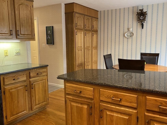 kitchen featuring dark stone countertops, brown cabinetry, baseboards, light wood-type flooring, and wallpapered walls