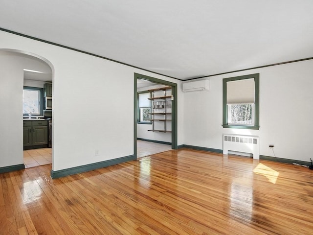 unfurnished living room with a healthy amount of sunlight, radiator heating unit, an AC wall unit, arched walkways, and light wood-style flooring