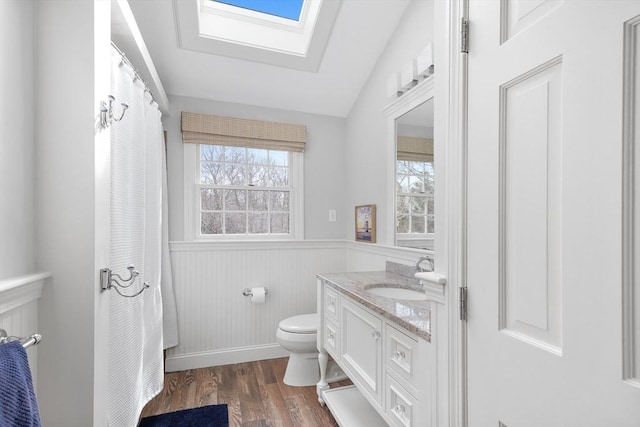 full bath with vanity, wood finished floors, a wainscoted wall, lofted ceiling with skylight, and toilet