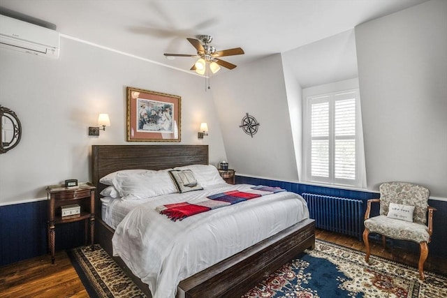 bedroom featuring a wall unit AC, radiator heating unit, wood finished floors, and ceiling fan