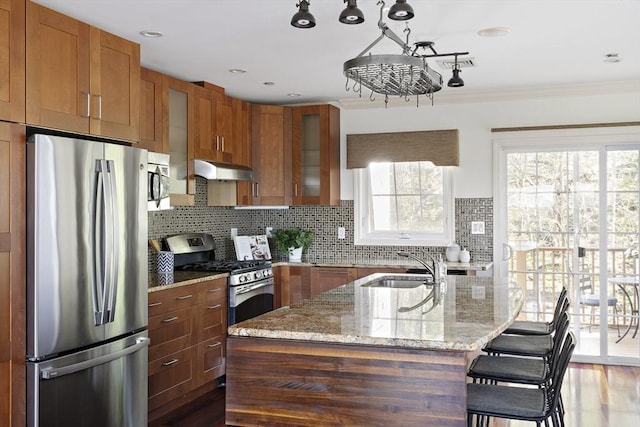 kitchen featuring light stone countertops, under cabinet range hood, brown cabinets, tasteful backsplash, and appliances with stainless steel finishes